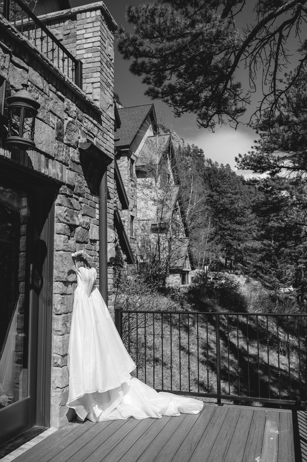 Black and white photo of a white wedding dress displayed on a balcony of a stone mansion surrounded by pine trees.