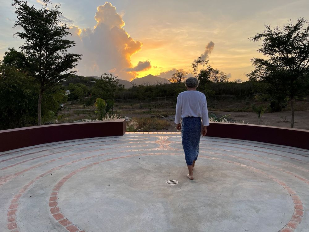 Person walking barefoot on circular platform at sunset with dramatic clouds and scenic landscape in background.