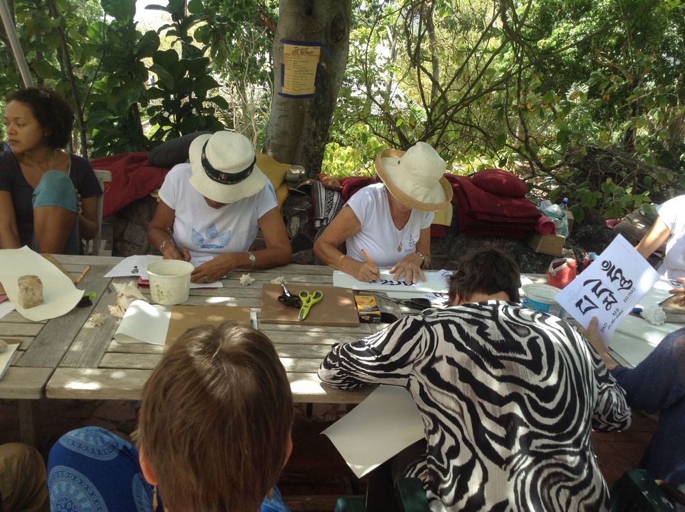 People in sun hats writing at an outdoor picnic table surrounded by lush green trees at a garden event.