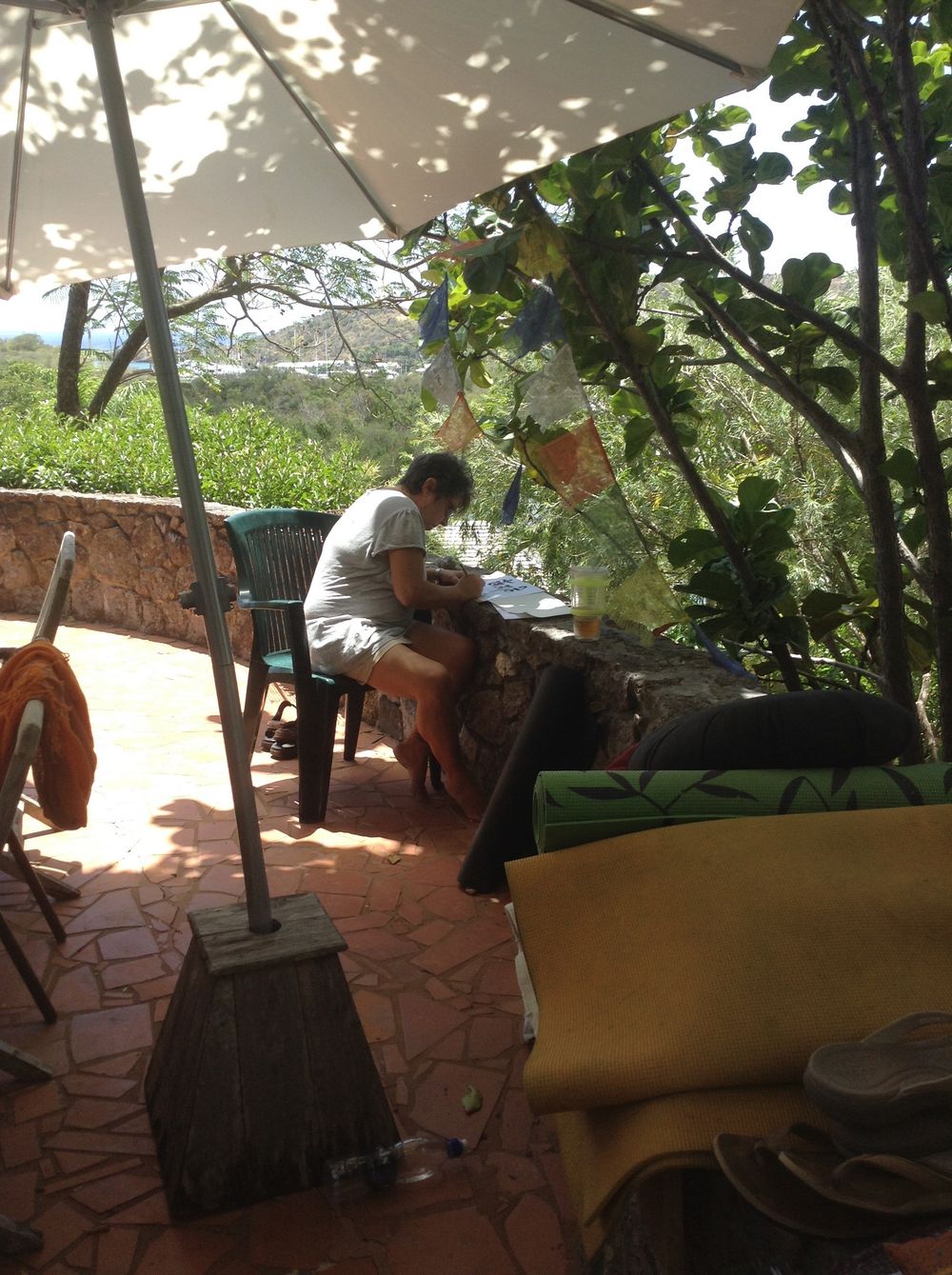 Person sitting under a white umbrella on a terracotta patio surrounded by lush green trees and plants.