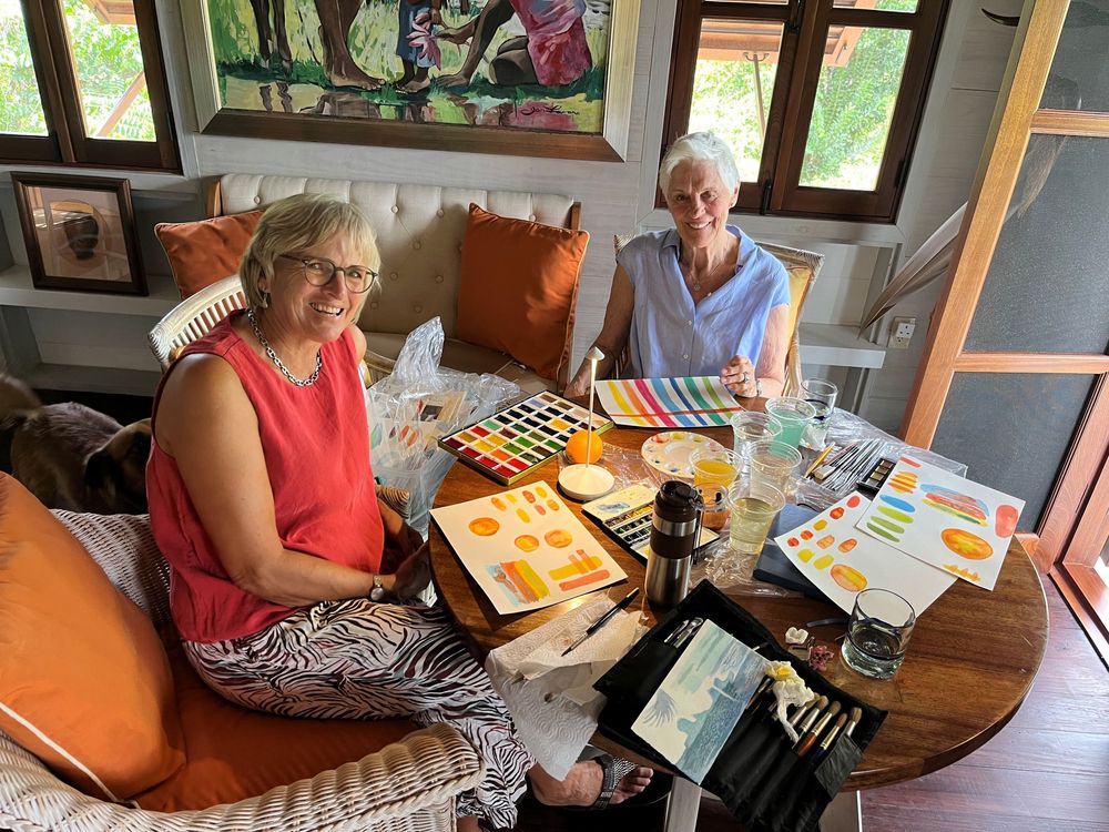 Two women smile while painting colorful artwork with watercolors at a wooden table in a bright, cozy art studio.