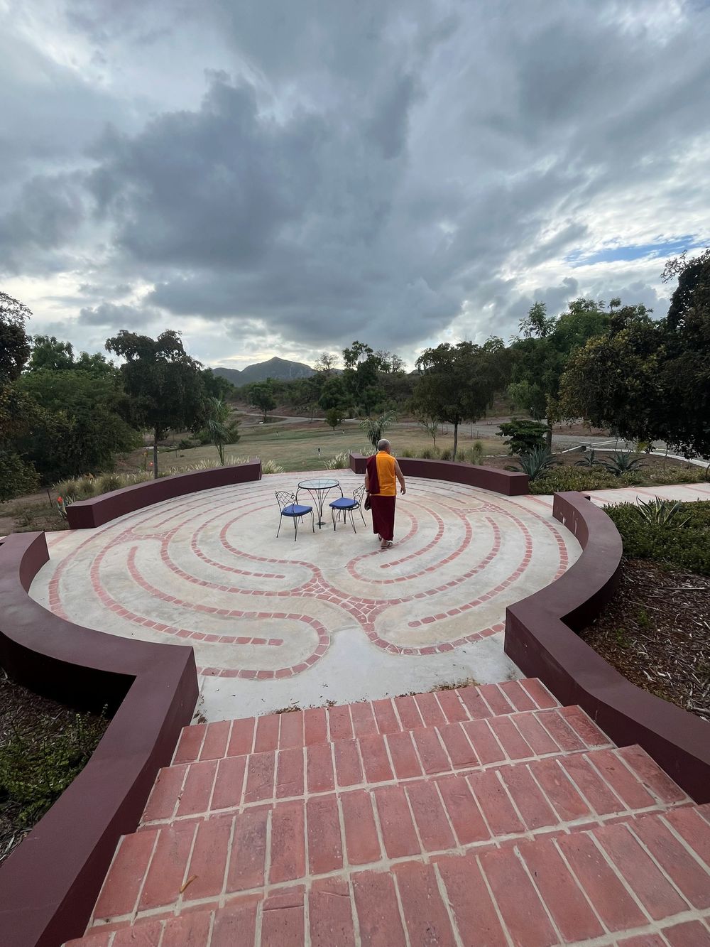 Person walking a brick labyrinth garden path under cloudy skies, surrounded by trees and arid landscape.