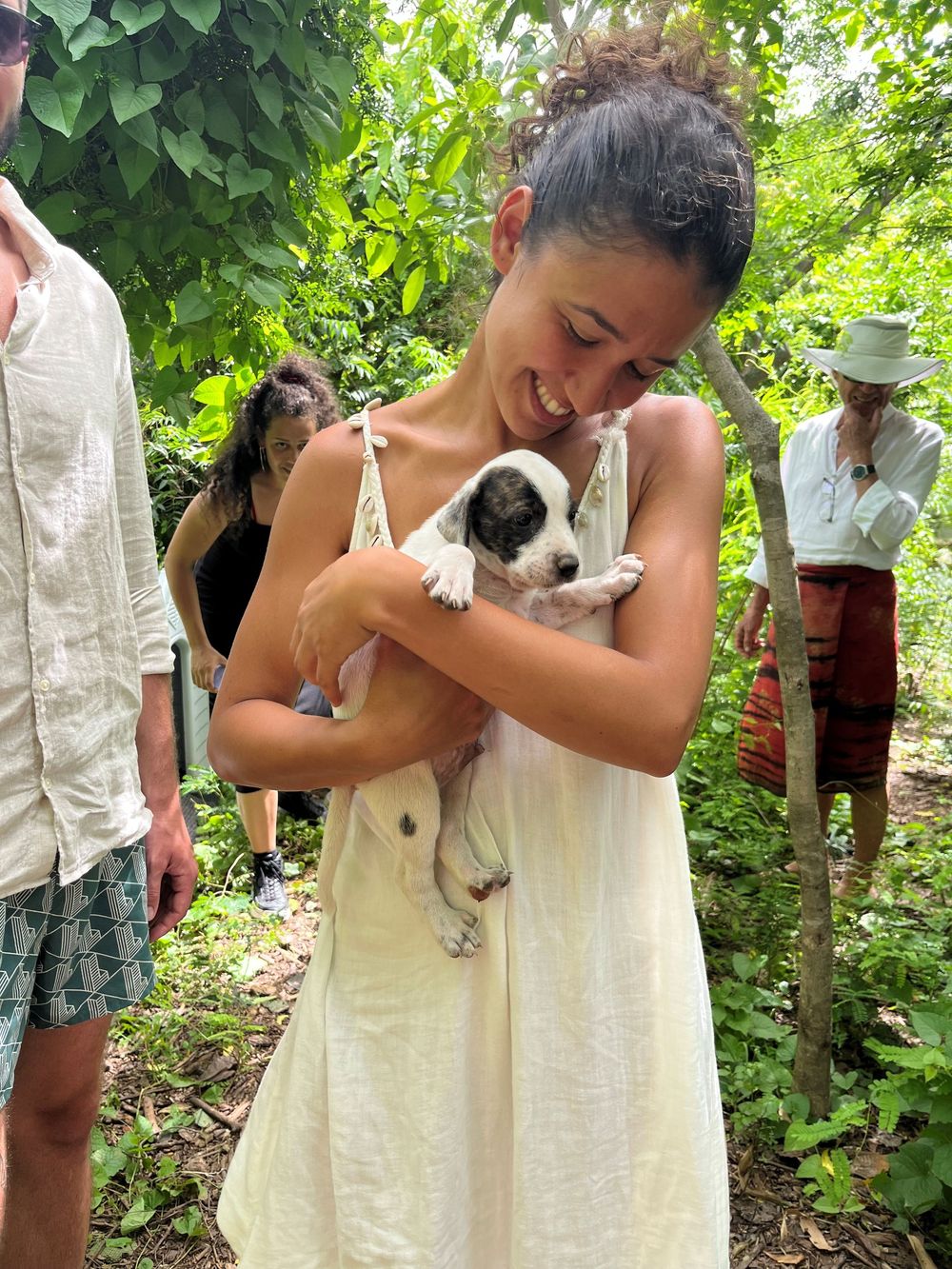Young woman in white dress smiling while holding a small black and white puppy outdoors among green trees.