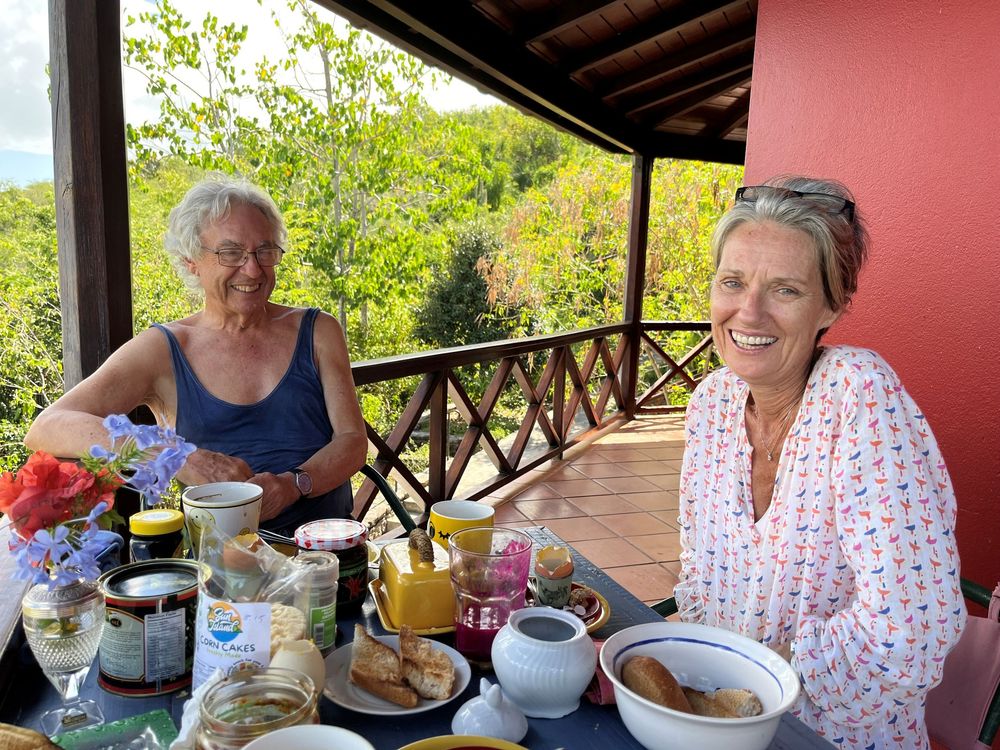 Two women smiling at an outdoor breakfast table on a porch with lush green trees in the background.