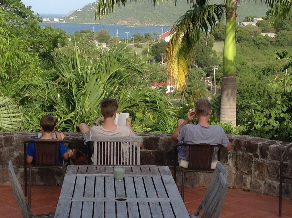 Three people sit on a tropical terrace with lush greenery and ocean views in the background.