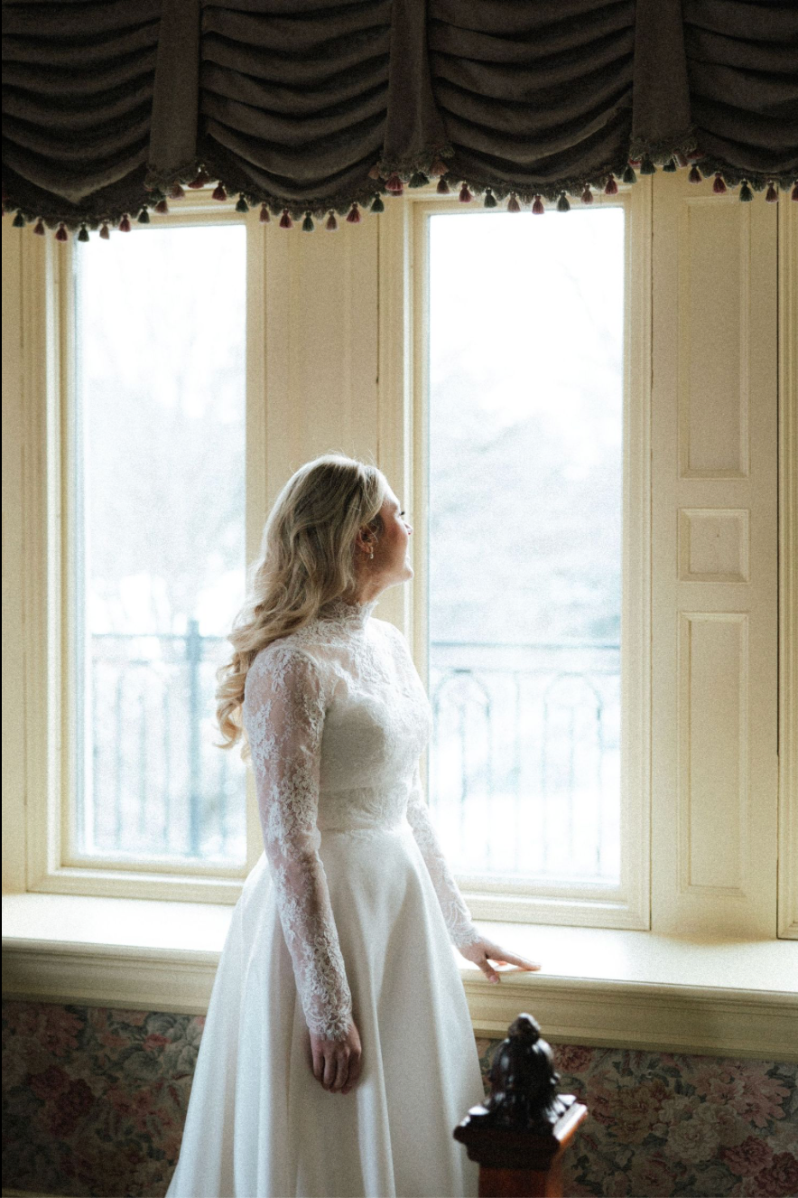 Bride in lace long-sleeve wedding gown gazing out a large window with elegant draped curtains in a classic interior.