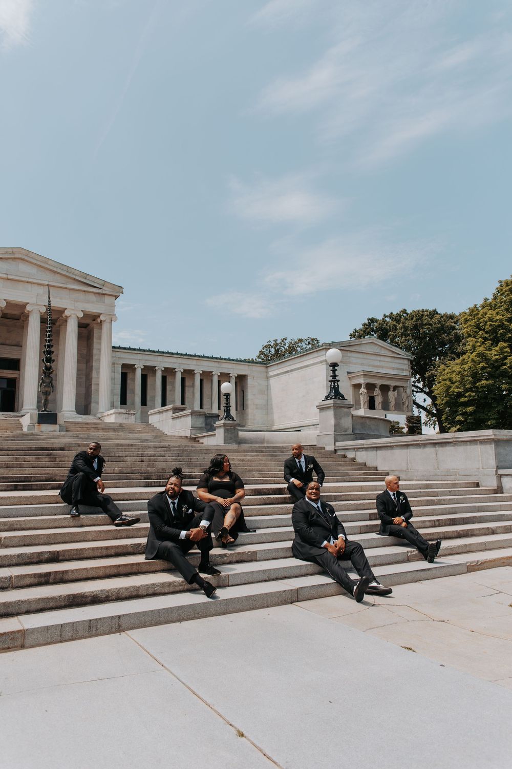 Group of men in black suits sitting on steps of a neoclassical building on a sunny day.