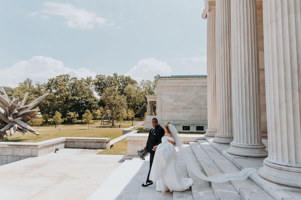 Bride and groom pose on grand neoclassical building steps with tall white columns and lush green trees in background.
