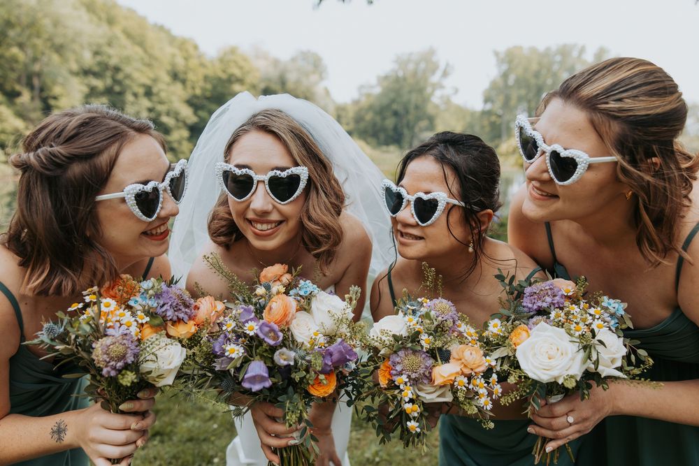 Bride and bridesmaids in green dresses wearing heart sunglasses, holding colorful floral bouquets outdoors.