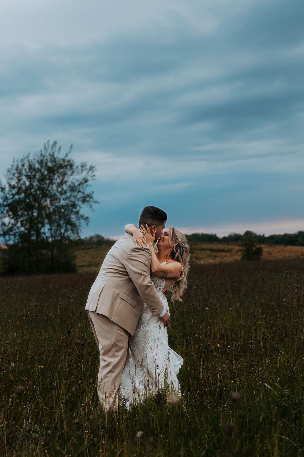 A couple shares a romantic kiss in a lush green field at dusk, with the groom in a tan suit and bride in a white gown.