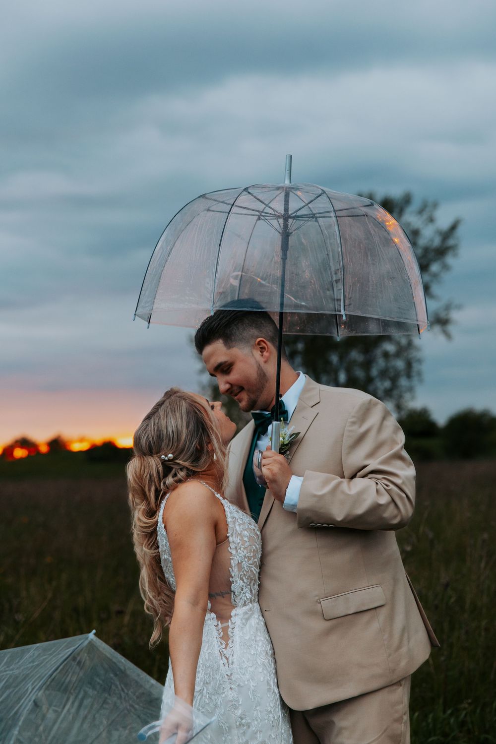 Bride and groom share a romantic moment under a clear bubble umbrella in a field at sunset.