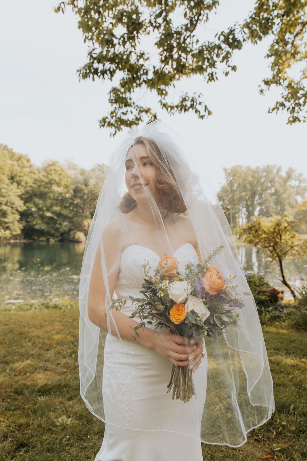 Bride in white strapless gown and veil holds colorful bouquet outdoors near a lake surrounded by lush green trees.