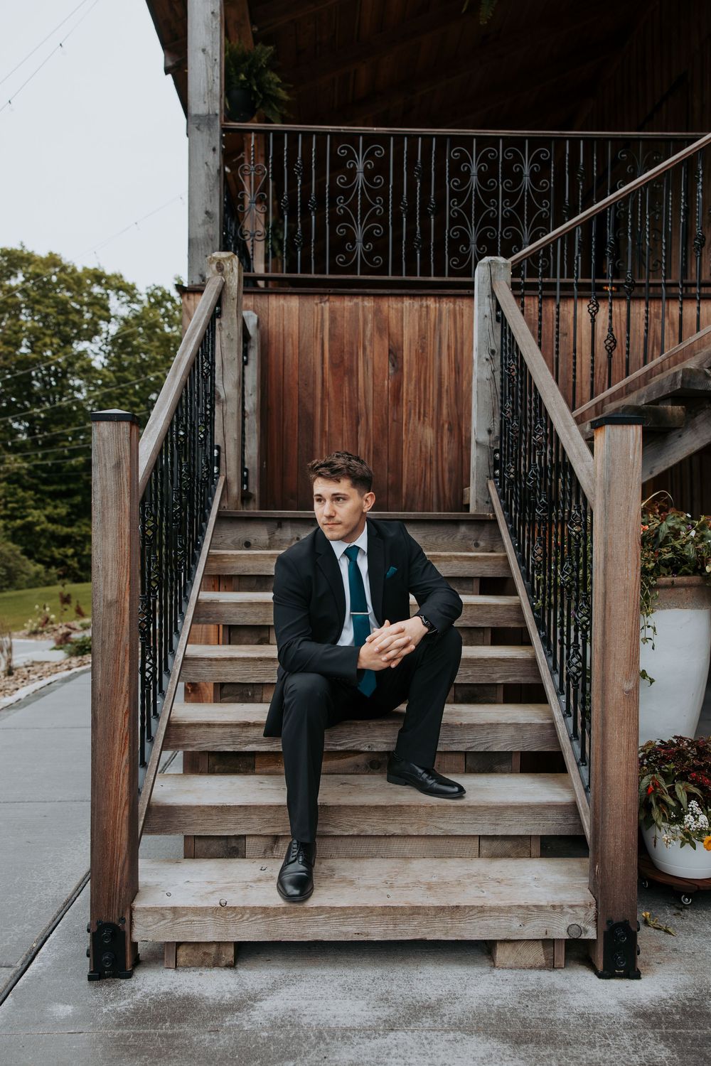 A well-dressed man in a dark suit sits on wooden outdoor stairs with ornate black iron railings.