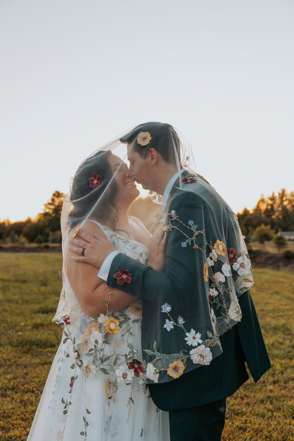 Bride and groom kissing under a floral embroidered veil in a sunlit field during their outdoor wedding ceremony.