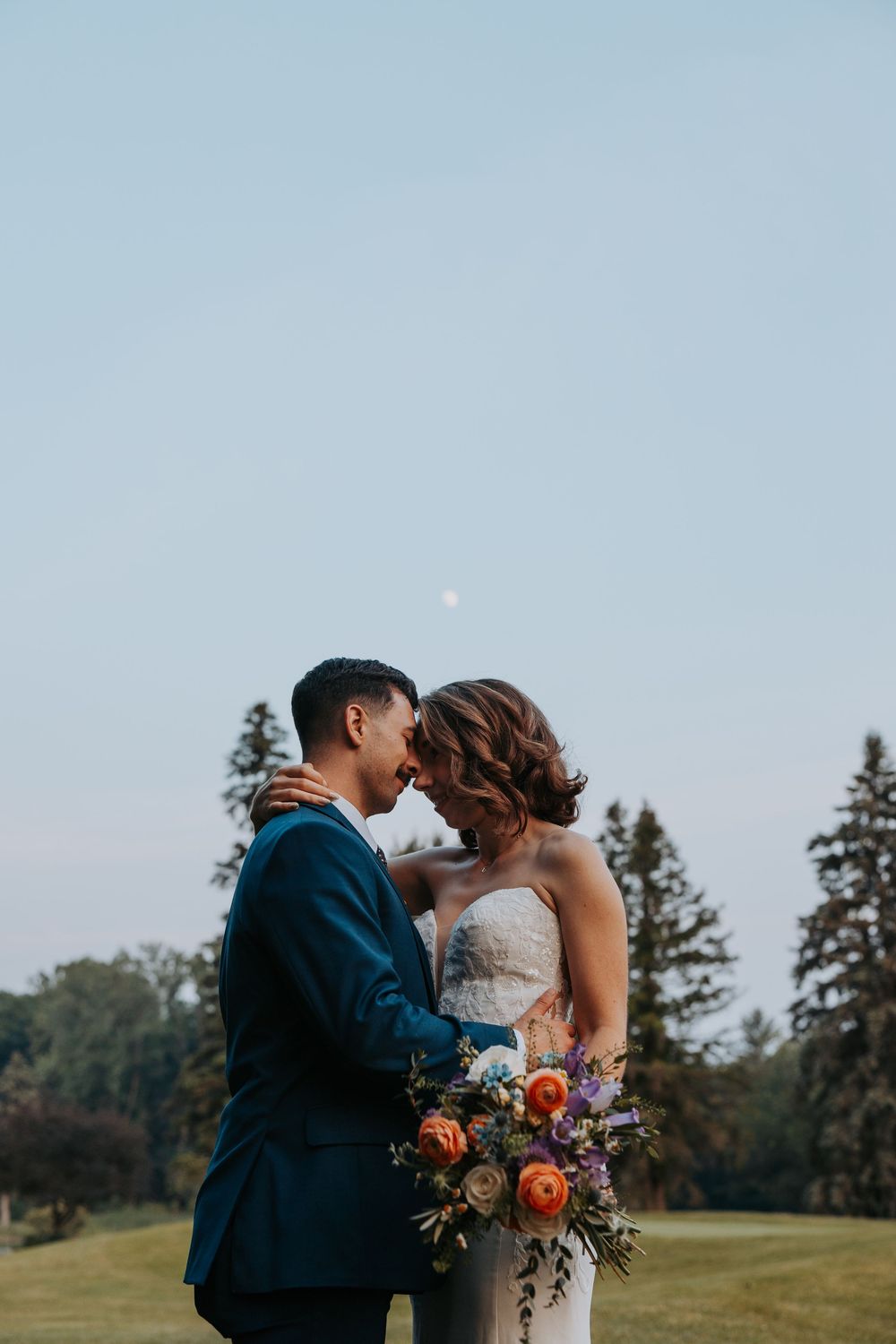 Bride and groom share a romantic moment outdoors, bride holding a vibrant bouquet of orange and purple flowers.