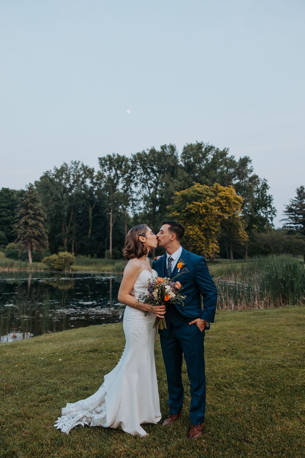 Bride and groom share a kiss outdoors near a pond at dusk, bride holding colorful bouquet, groom in navy suit.