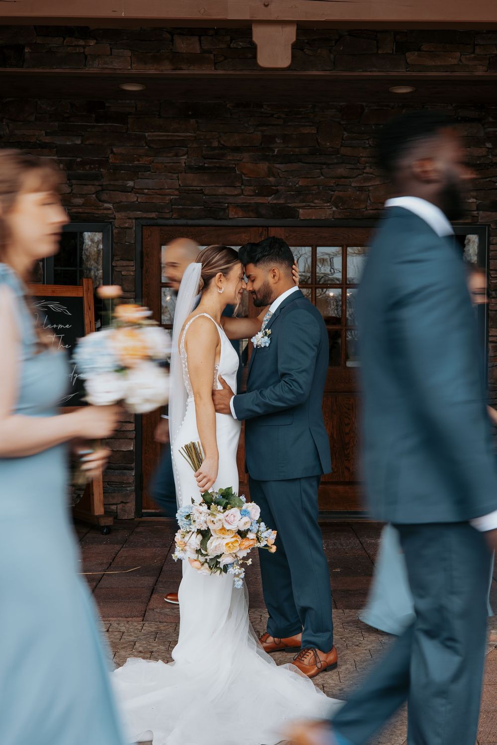Bride and groom share an intimate moment, surrounded by wedding party in blue attire, outside a stone building.