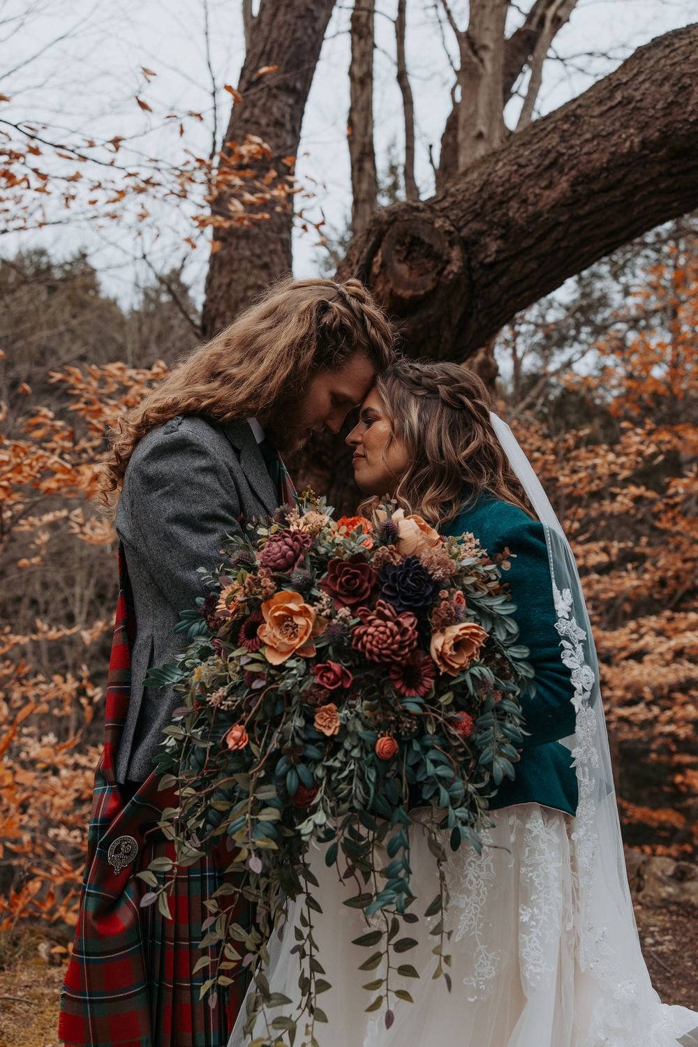 Romantic autumn wedding couple touching foreheads holding lush burgundy and orange floral bouquet in forest setting.