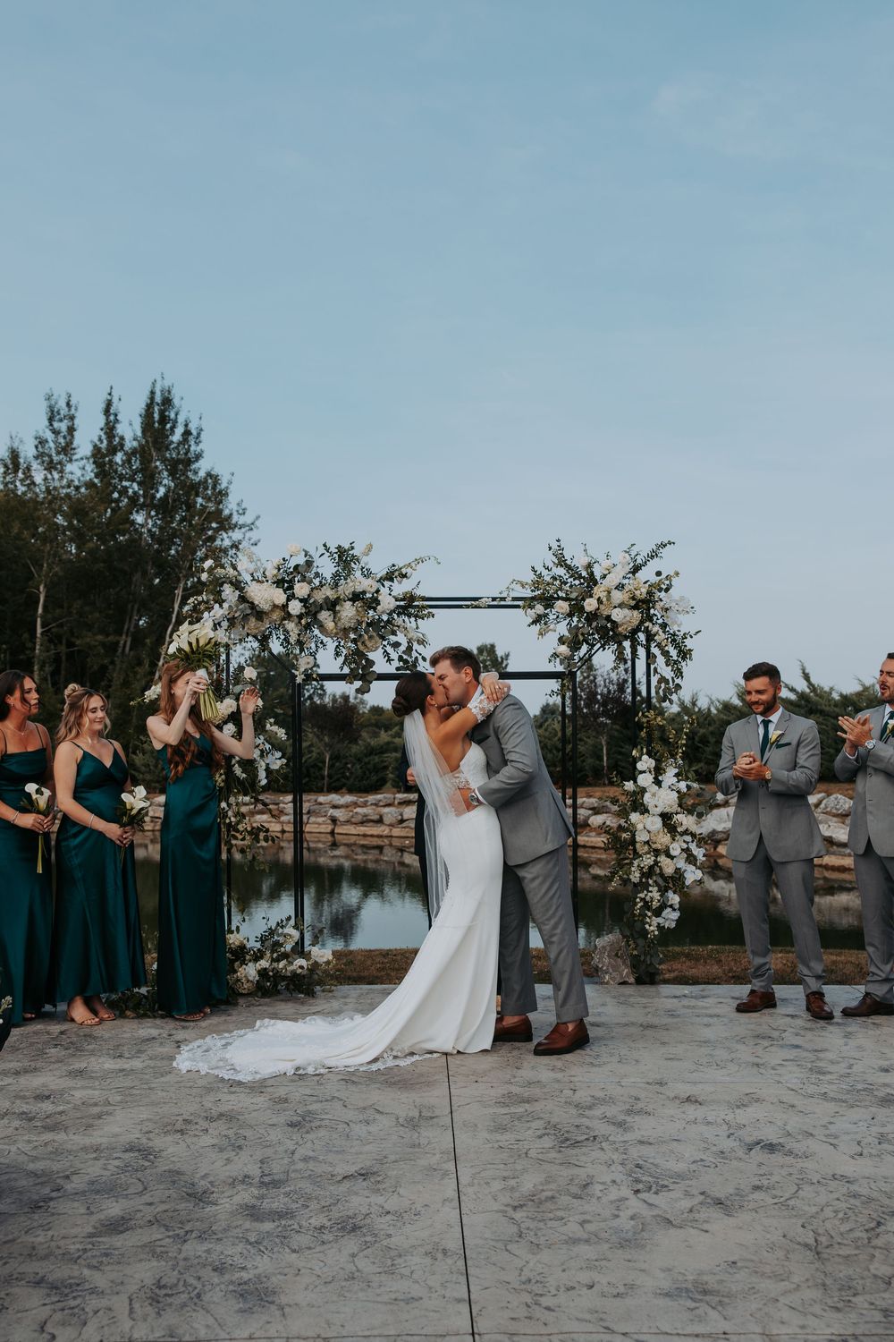 Bride and groom share first kiss at outdoor wedding ceremony with floral arch, bridesmaids in emerald green dresses.