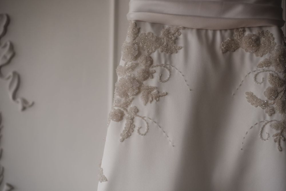Elegant white lace wedding dress hanging against a light colored wall with ornate molding details.