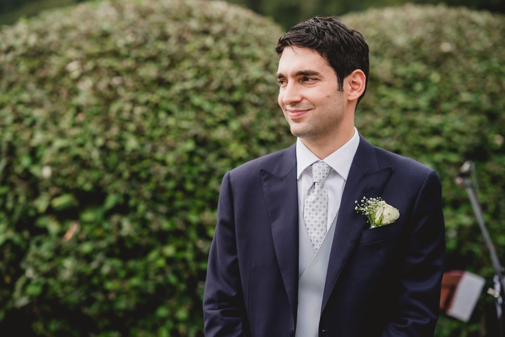 Groom in navy suit with light tie standing in front of lush green garden backdrop.