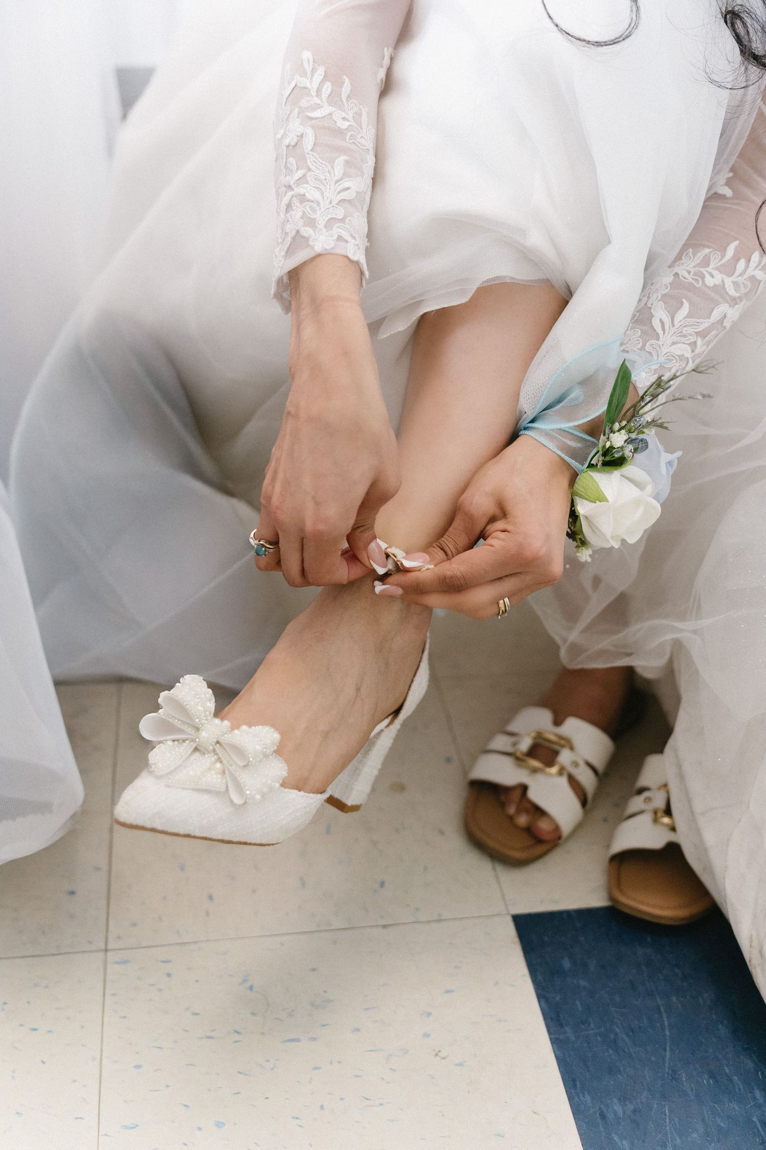 Bride being helped into white floral heels by bridesmaid wearing wrist corsage on wedding day.