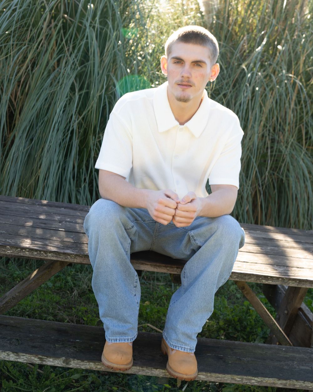 Young man in white polo shirt and jeans sitting on wooden bench outdoors with tall grass background.