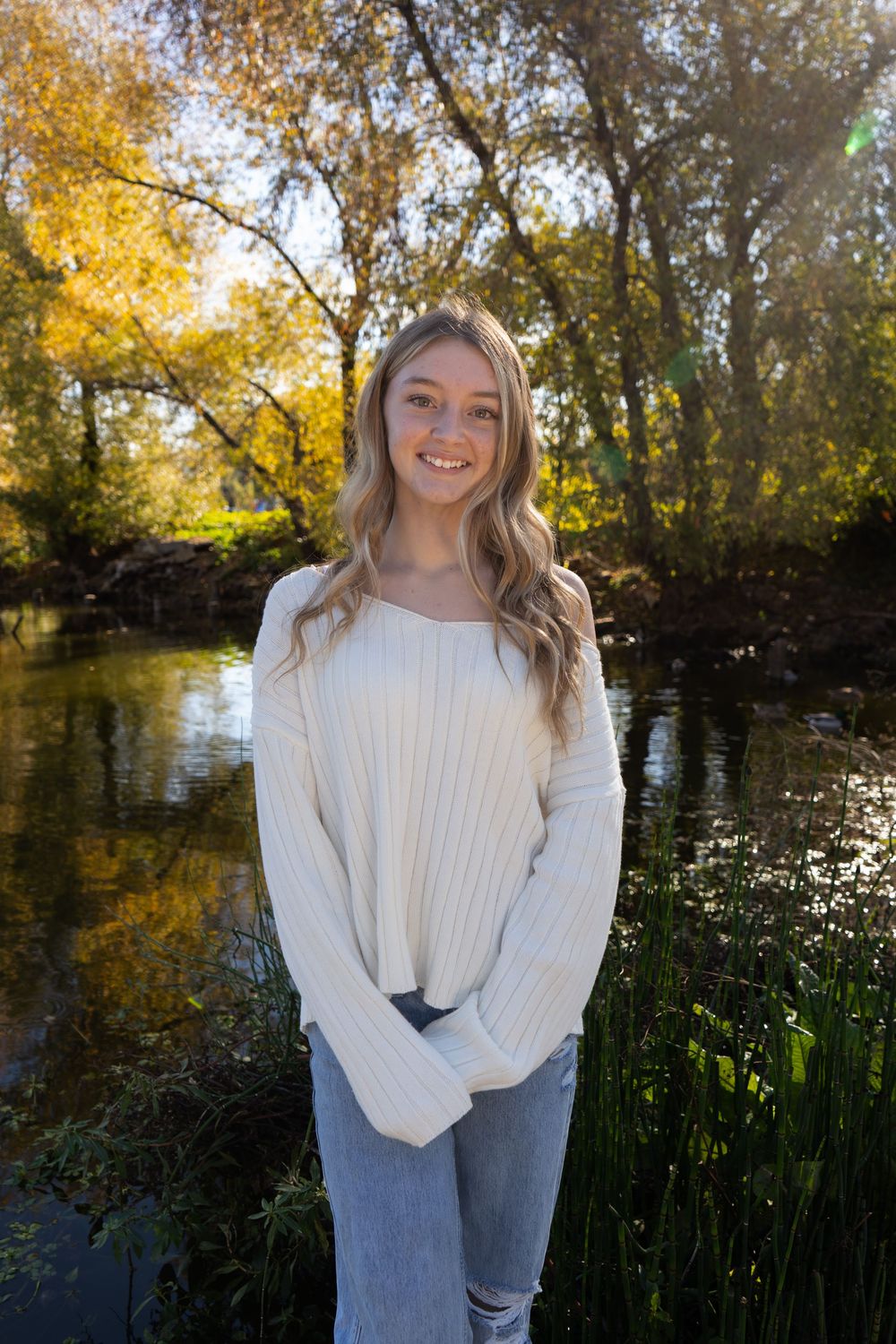 Smiling young woman wearing a white ribbed sweater and jeans standing near a sunlit forest creek.
