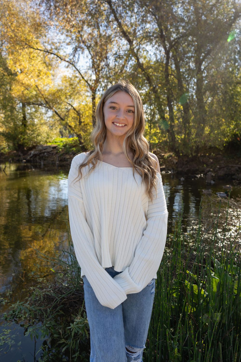 Young woman in cream sweater and jeans smiling outdoors by a serene creek surrounded by autumn trees.