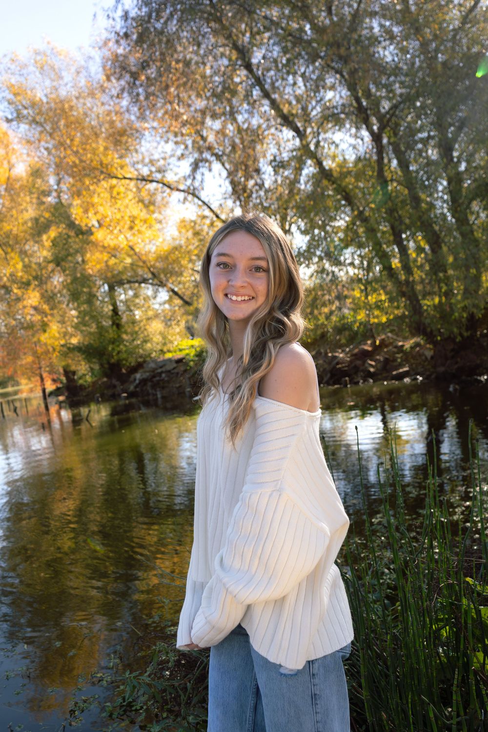 Young woman in white off-shoulder sweater smiling by a sunlit river surrounded by autumn trees.