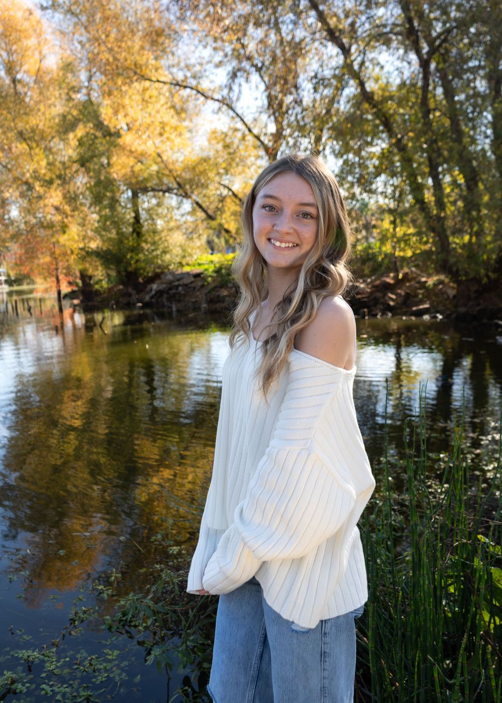 Smiling young woman in white off-shoulder sweater and jeans posing by a serene tree-lined pond in autumn.