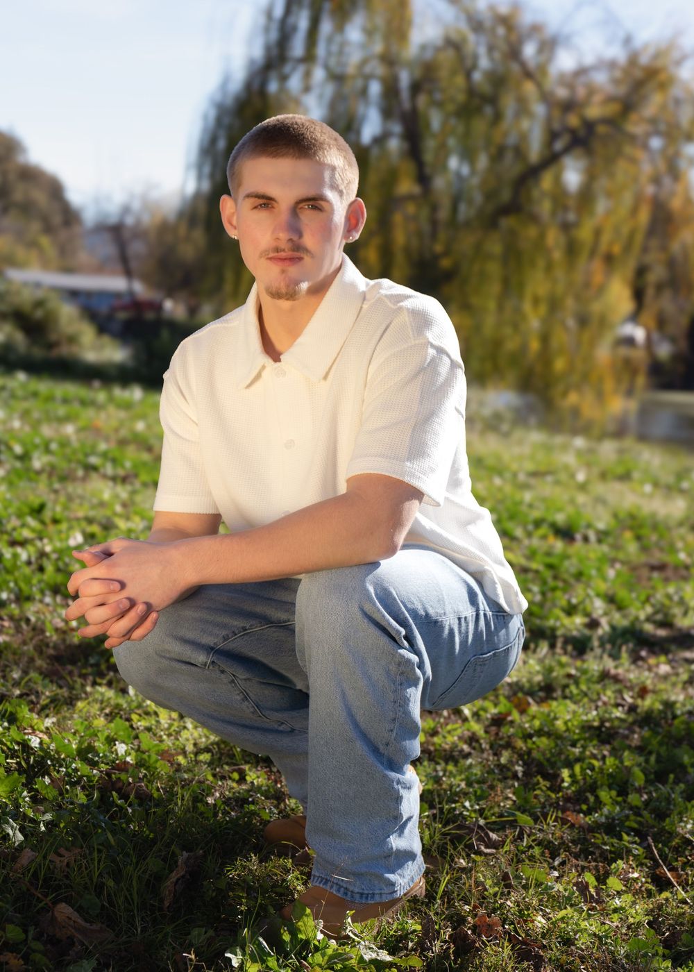 Young man in white polo shirt and jeans crouching outdoors on green grass with trees in background.