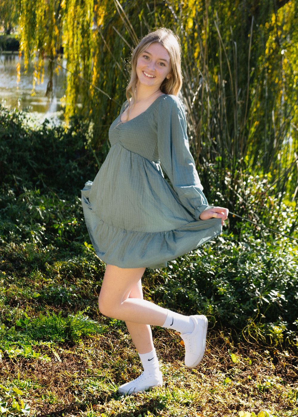 Young woman in sage green tiered mini dress and white Nike socks poses outdoors near willow tree on sunny day.
