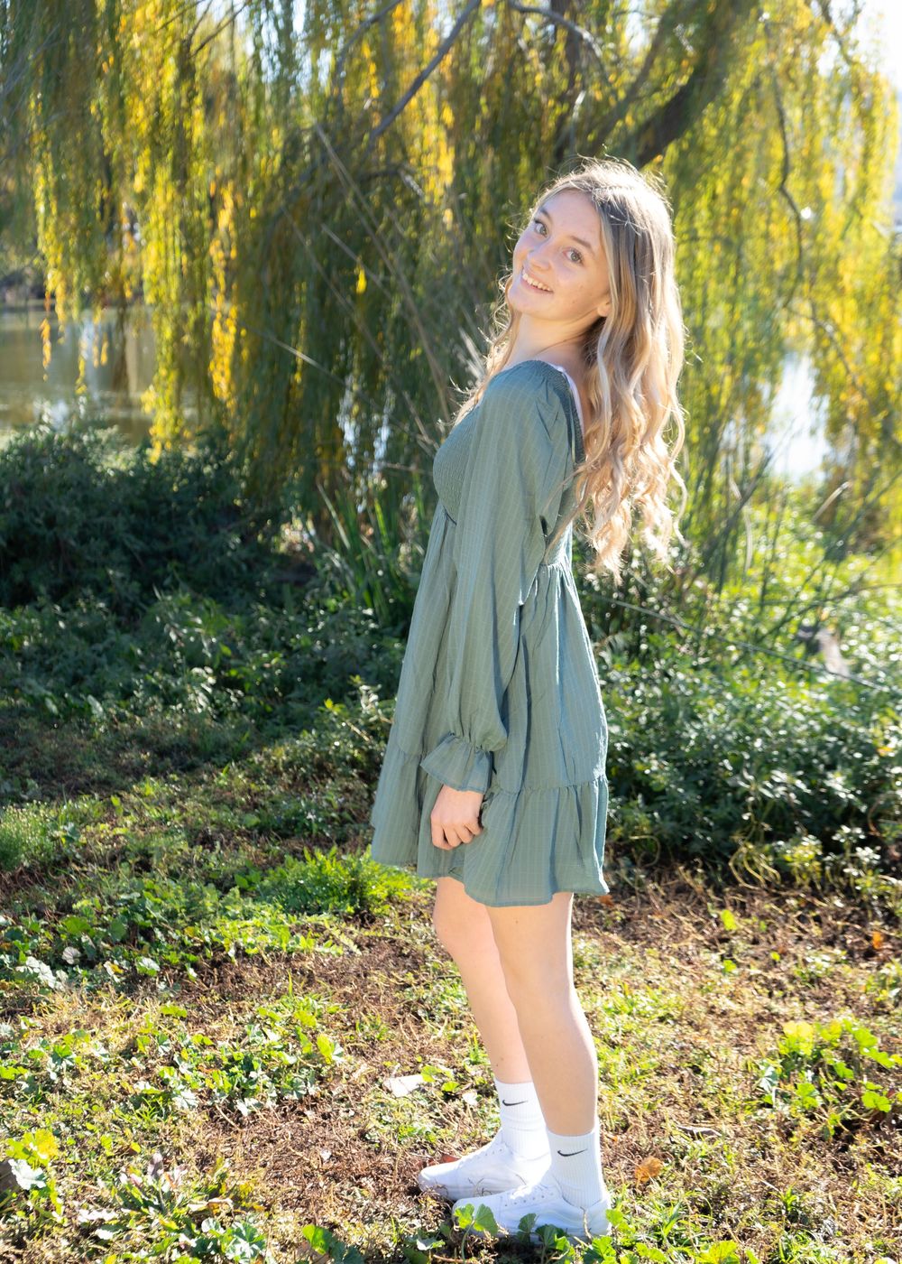 Young woman in sage green dress and white socks smiling outdoors near a weeping willow tree on a sunny day.
