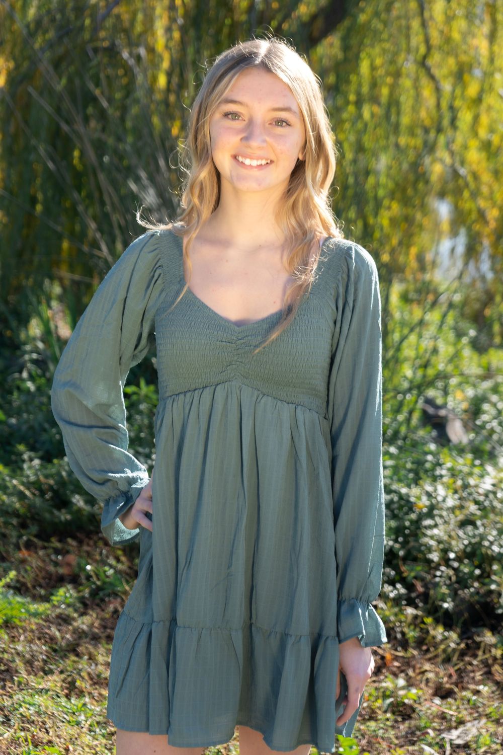 Young woman smiling in olive green long-sleeve babydoll dress, posing outdoors among lush greenery.