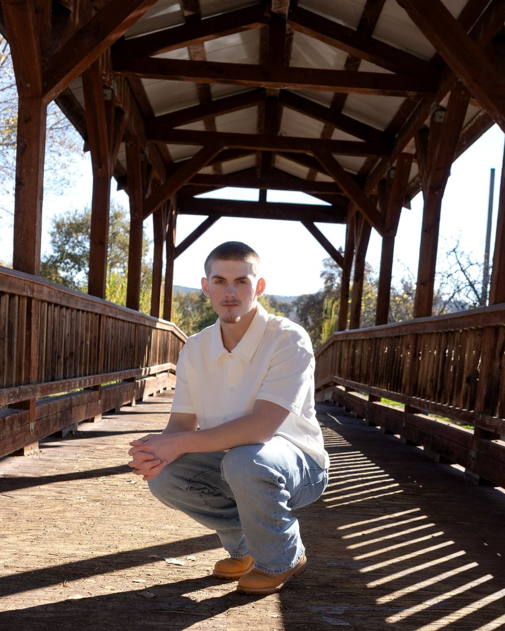 Young man in white polo crouching on a wooden covered bridge pathway in a sunny outdoor setting.