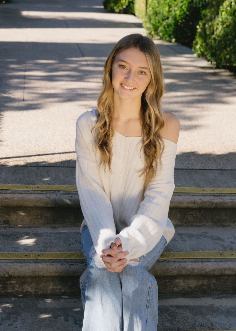 Young woman with long wavy hair smiling, wearing off-shoulder white top and jeans, sitting on outdoor steps in sunlight.