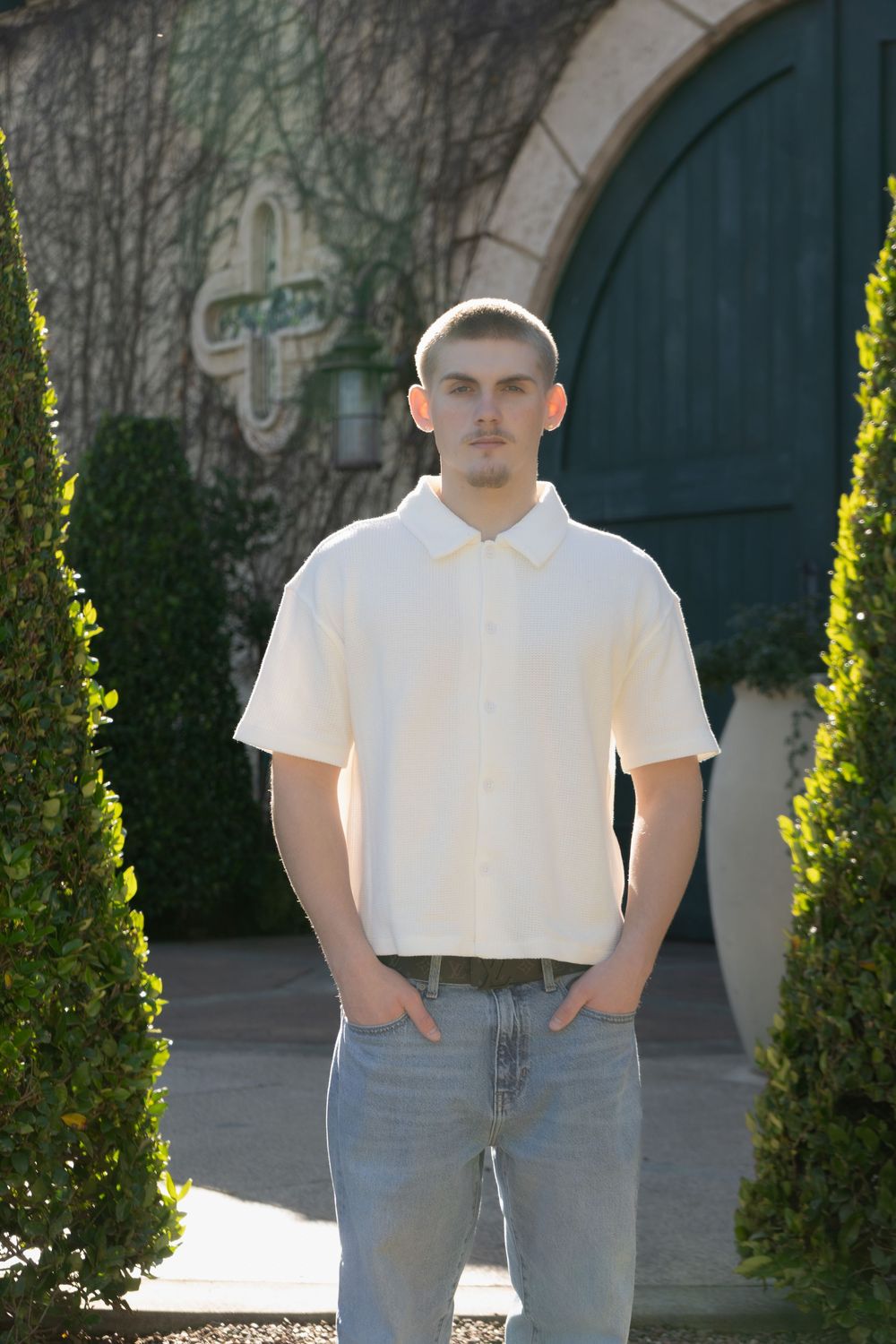 Young man in cream shirt and jeans standing between trimmed hedges outside a building with green doors.