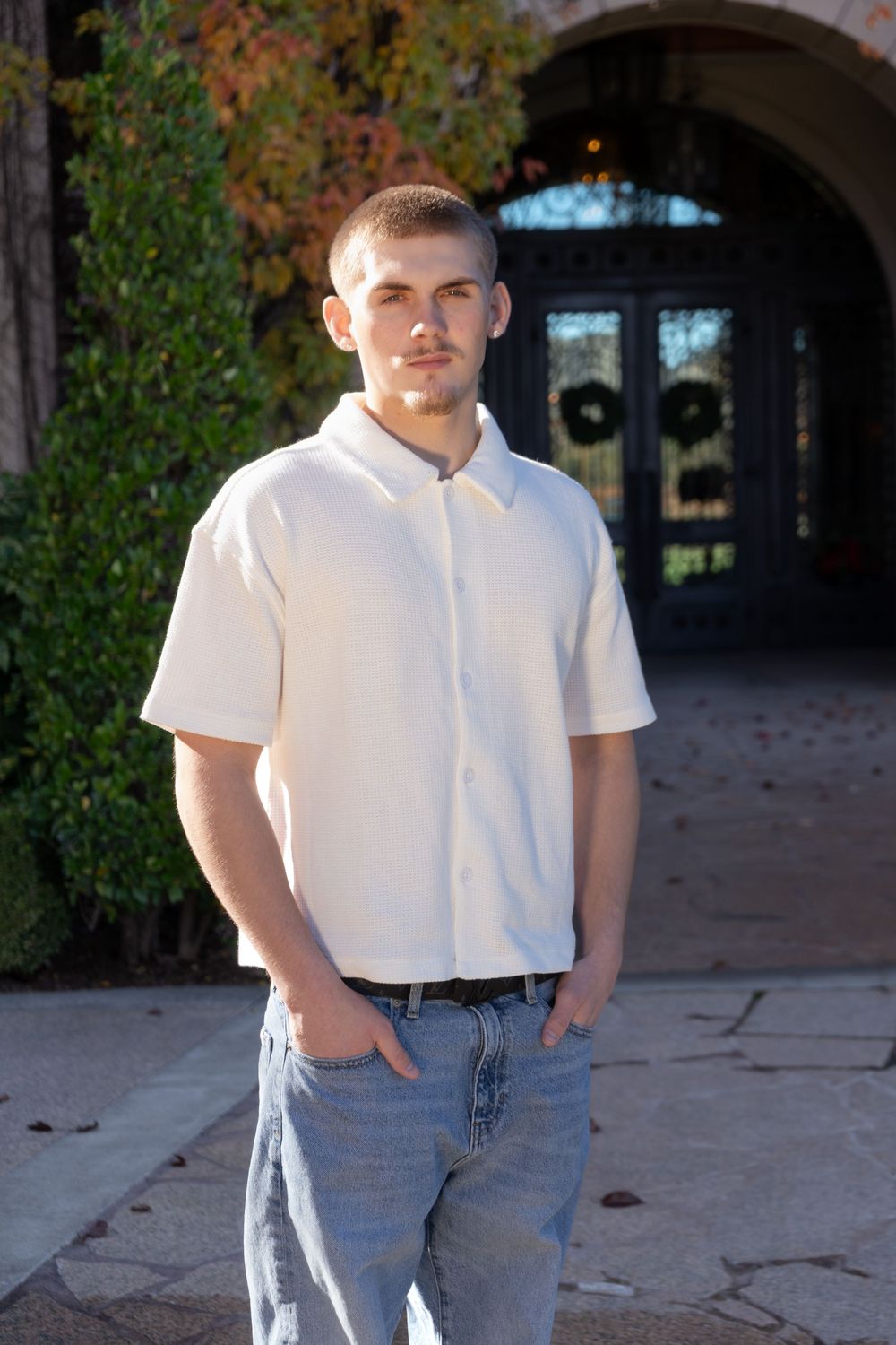 Young man in cream button-up shirt and jeans posing outdoors near an arched doorway.