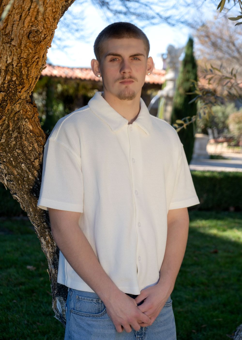 Young man in white short-sleeve button-up shirt and jeans standing outdoors near a tree.