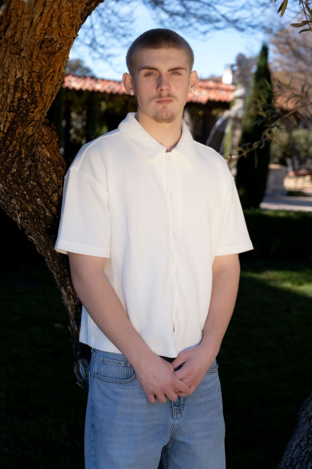 Young man in white polo shirt and jeans standing outdoors near a tree with a building in background.