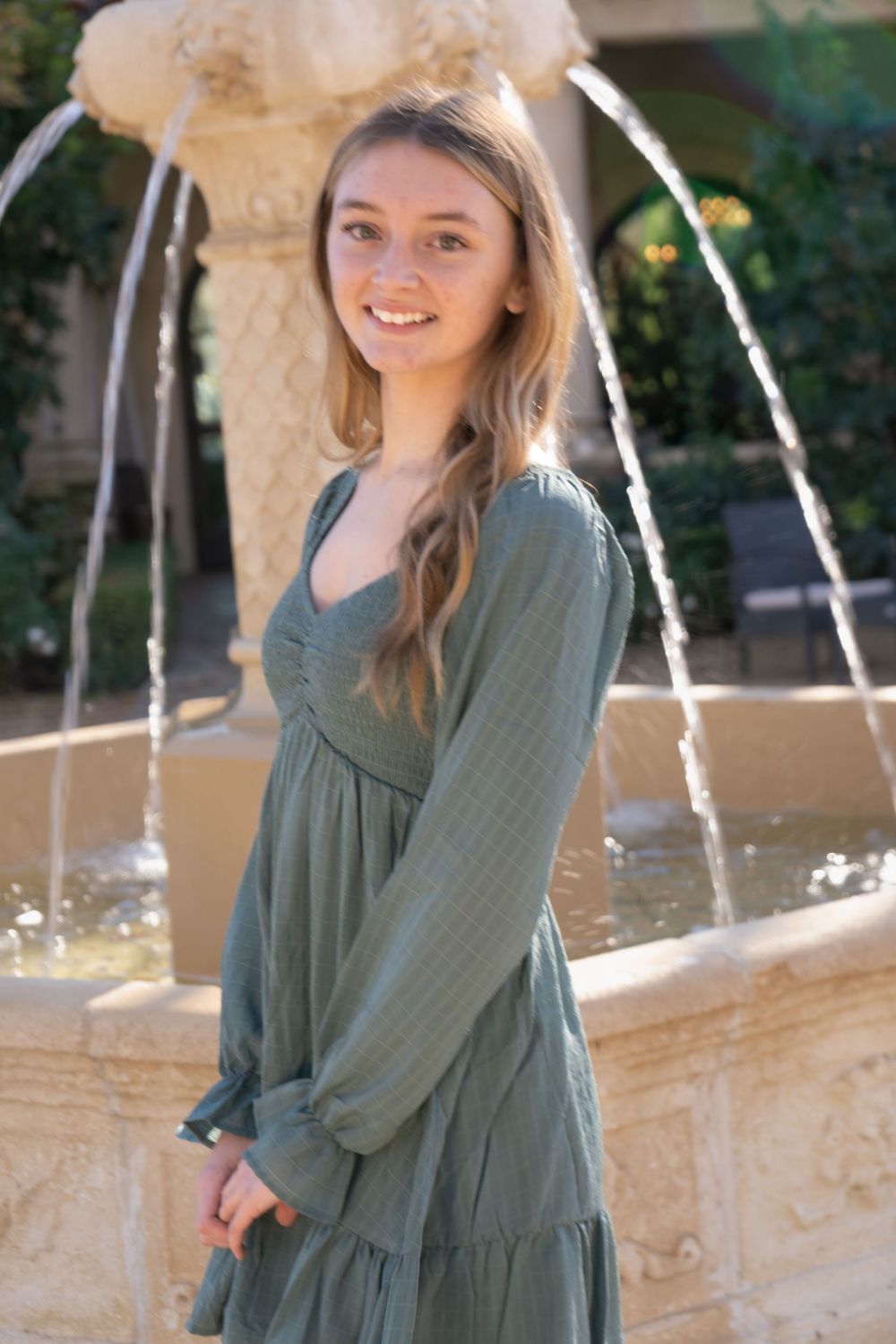 Smiling young woman in sage green tiered babydoll dress standing by stone fountain outdoors.