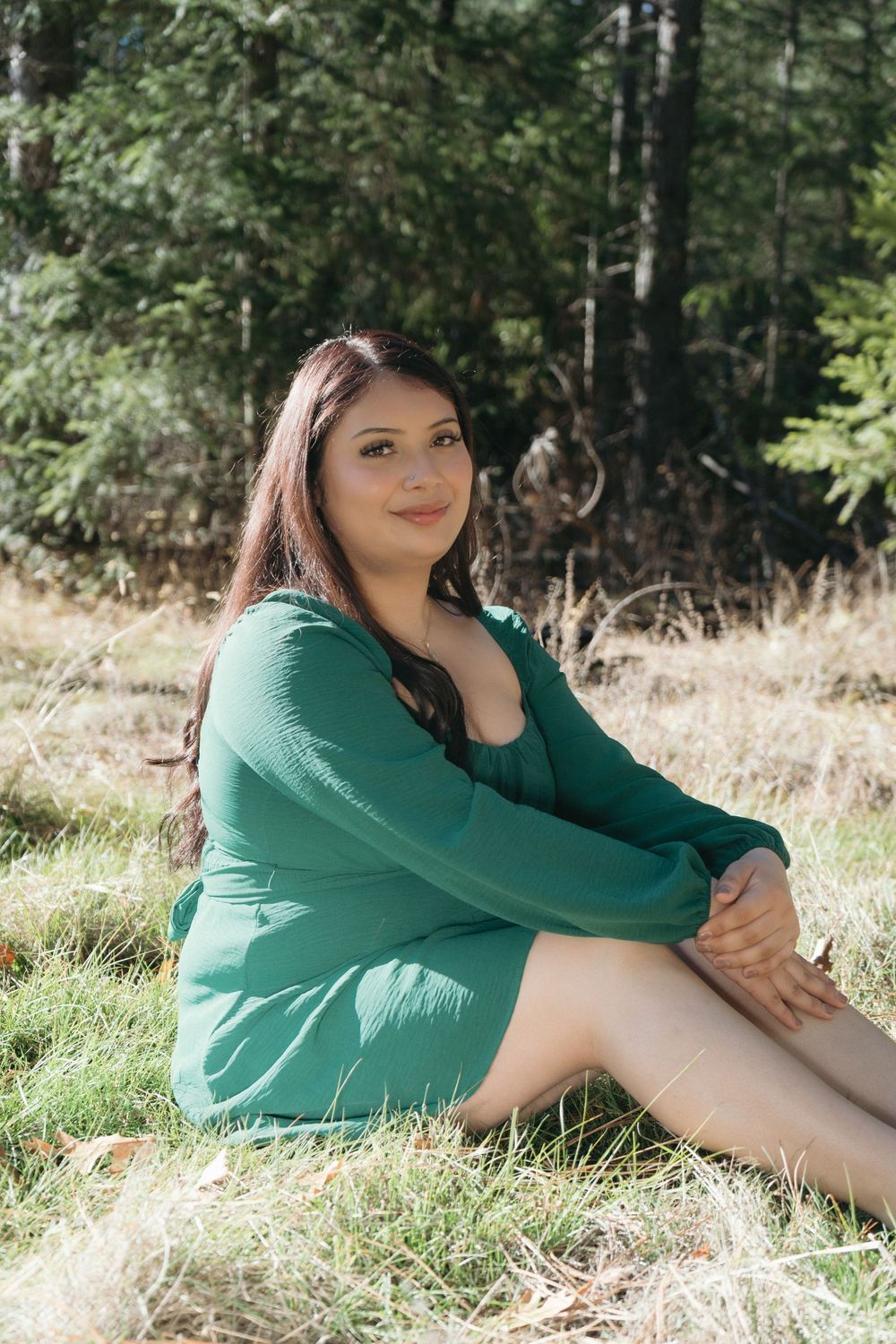 Young woman in green dress sitting on grass in front of forest, smiling at camera on sunny day.