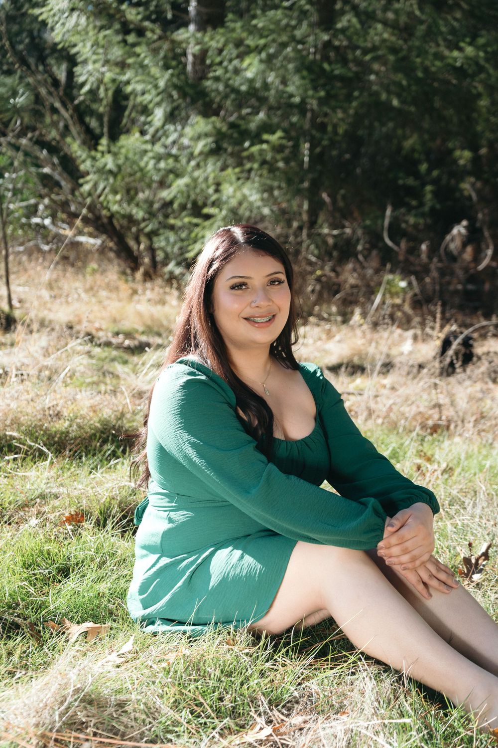 Young woman in green dress sitting on grass outdoors, smiling at camera with trees in background.
