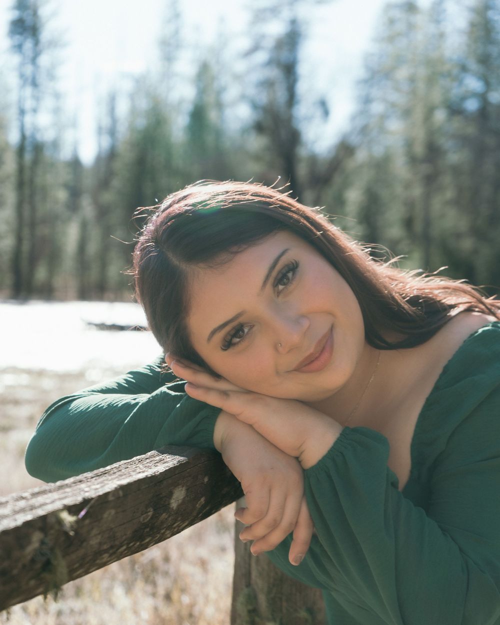 Young woman with brown hair smiling outdoors, leaning on a wooden fence with snowy forest background.