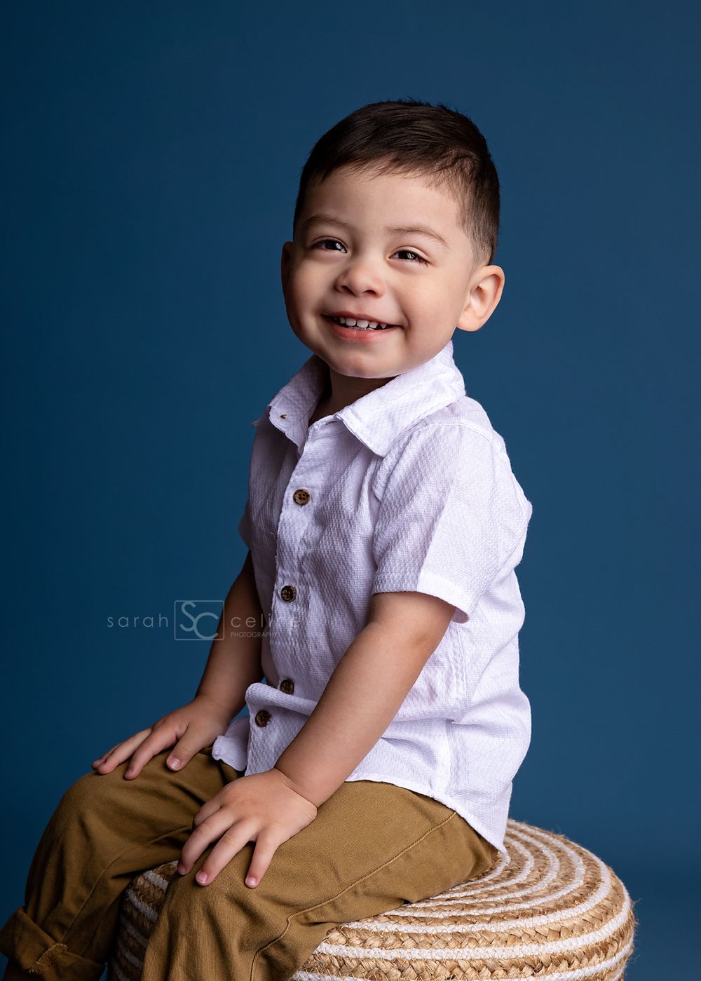 A smiling toddler boy in a white button-up shirt and tan pants sits on a woven stool against a blue backdrop.