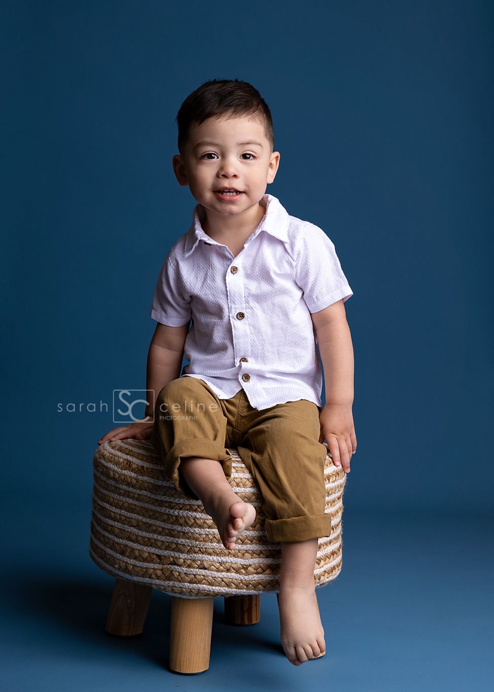 A young boy in a white shirt and tan pants sits barefoot on a woven stool against a blue background.