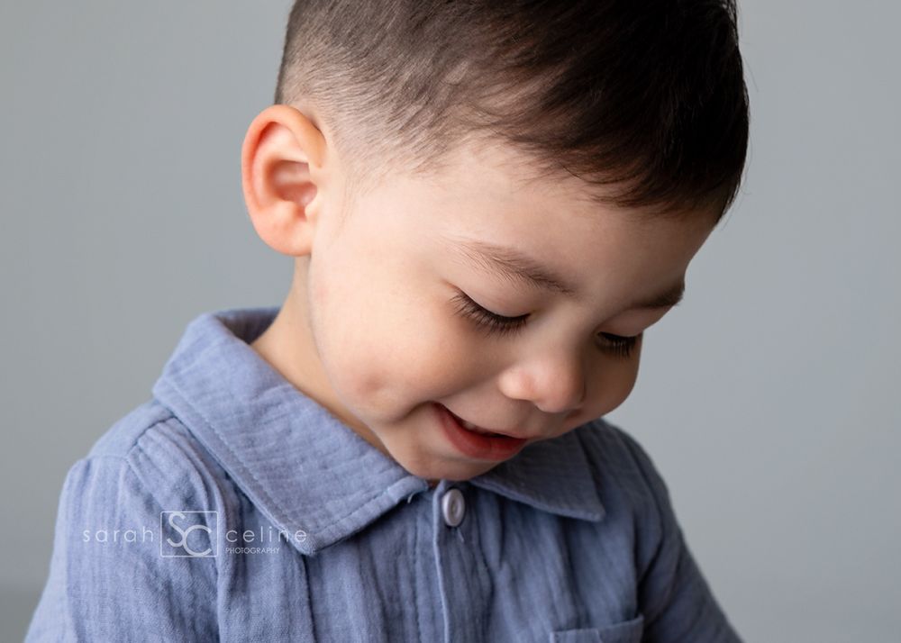 A young toddler boy with a short haircut smiles downward, wearing a blue chambray shirt against a gray background.
