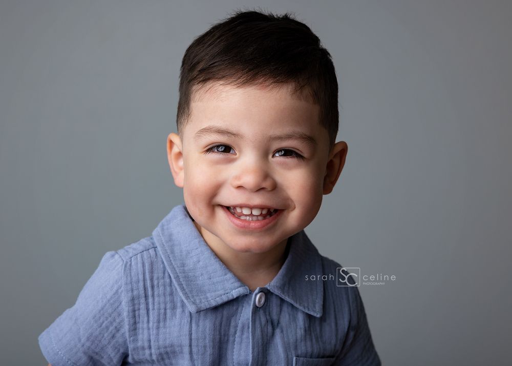 Smiling young boy with dark hair wearing a blue shirt, photographed against a gray studio background.