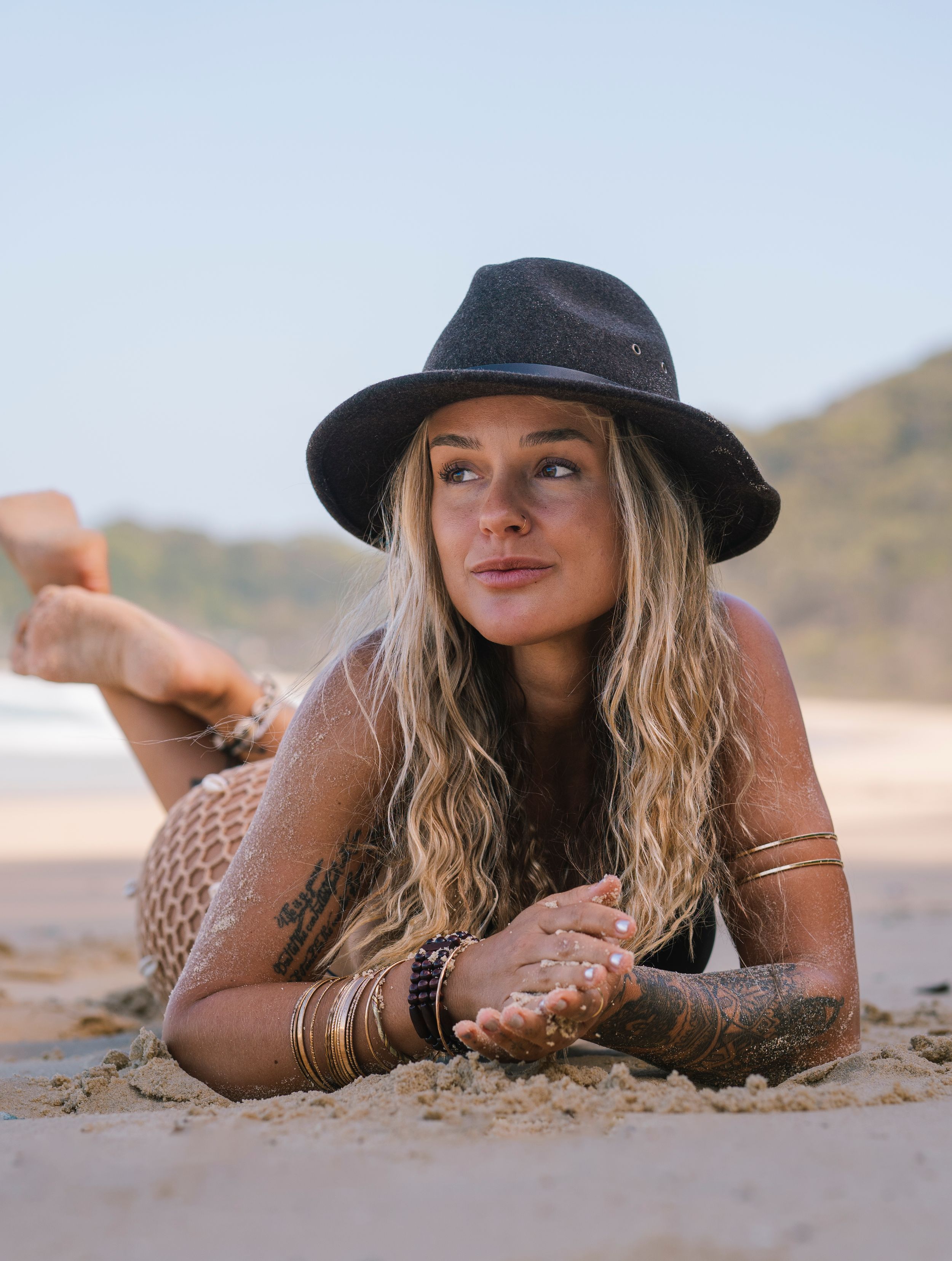 Blonde woman in black hat lying on sandy beach with gold jewelry and tattoos.